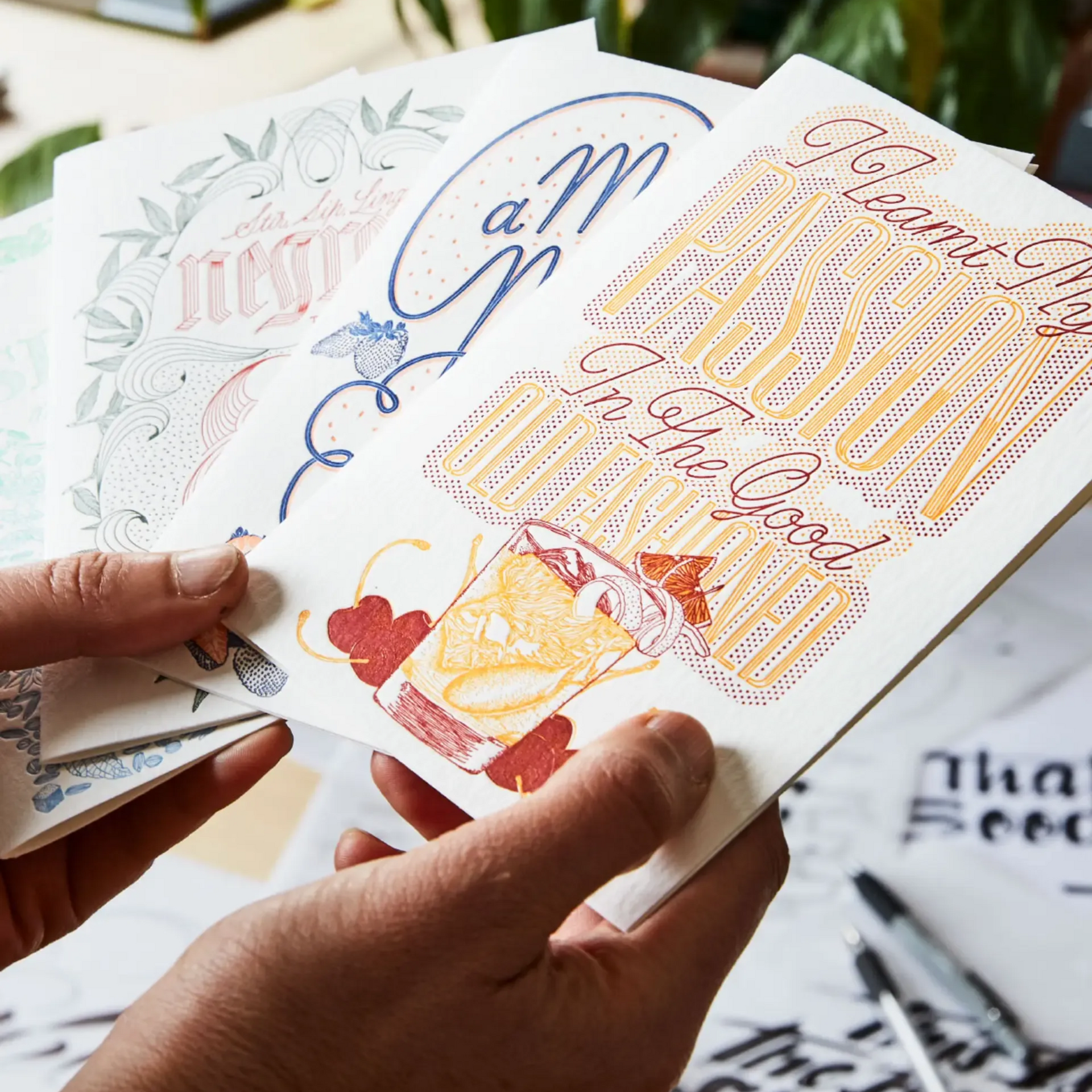 Hands holding several illustrated greeting cards with intricate calligraphy and cocktail art on a white desk.