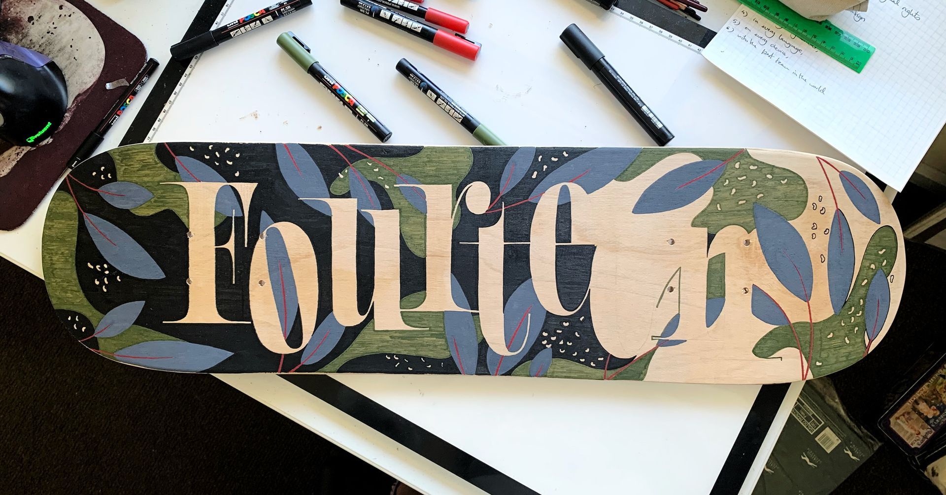 A decorated skateboard deck with a leafy pattern in green and blue, displaying the word 