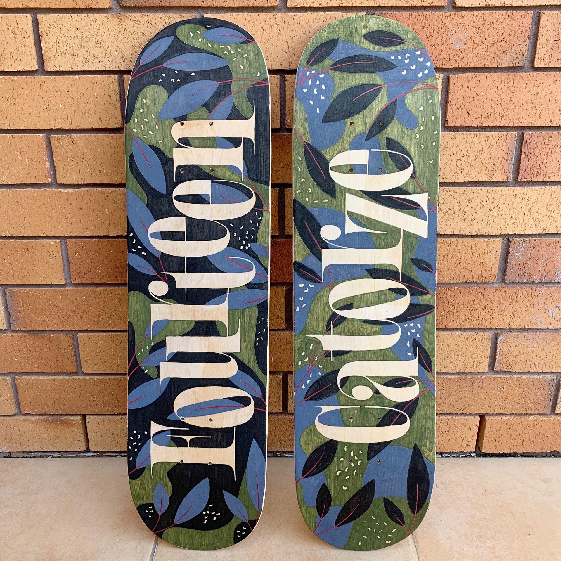 Two custom-painted skateboards with leaf patterns in blue and green, leaning against a brown brick wall.
