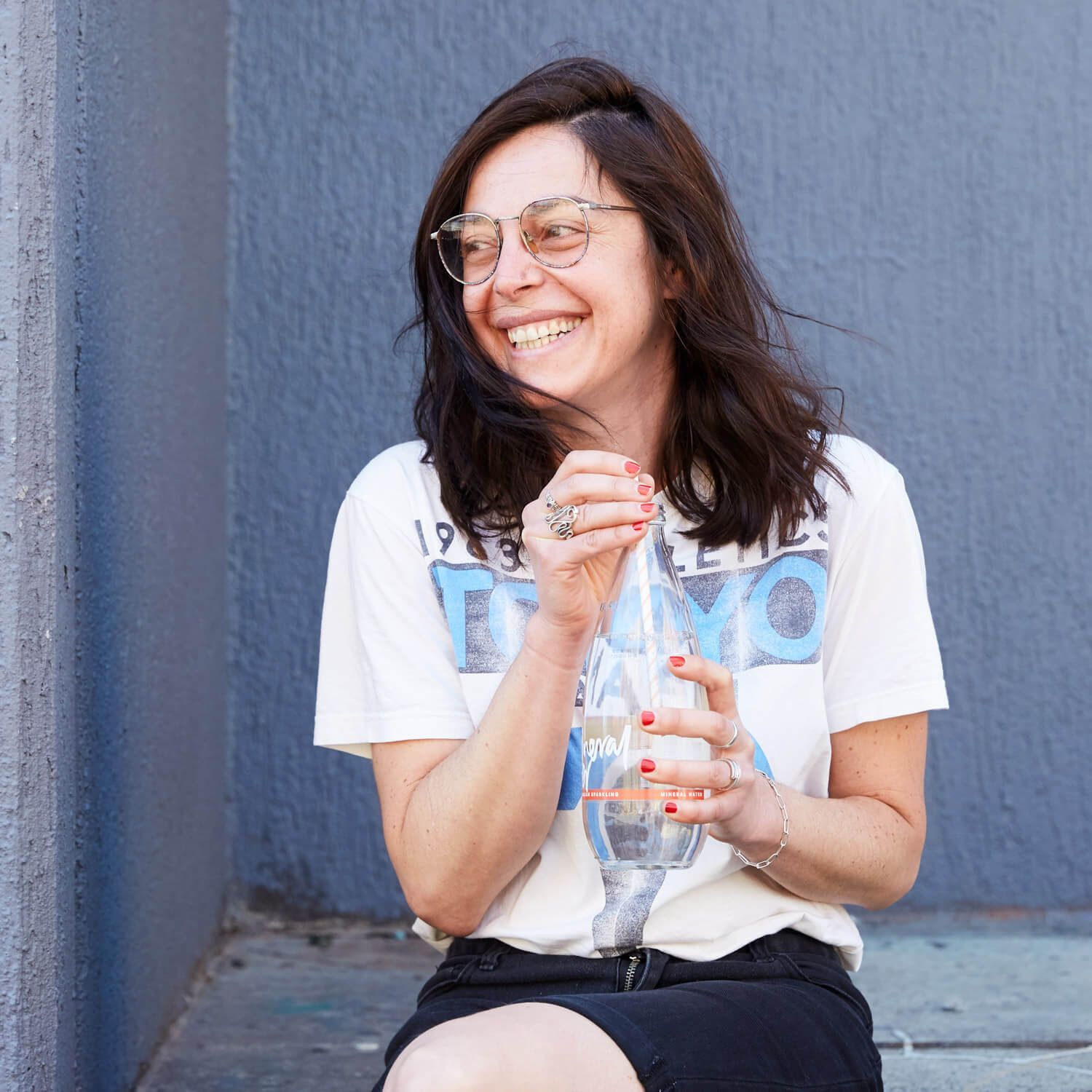 Maria Montes smiling in glasses holding a clear bottle, sitting against a blue wall