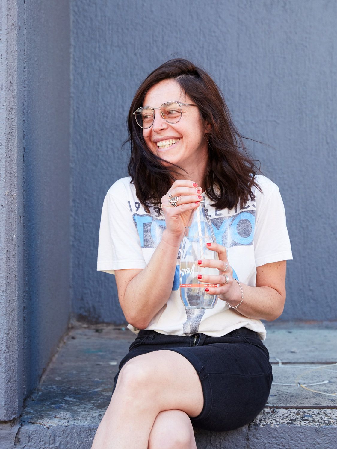 Woman sitting by a wall, smiling and holding a water bottle.