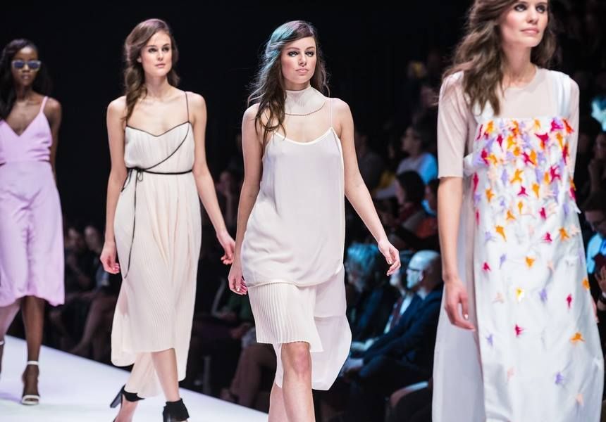 Fashion models walking a runway in pale dresses, one with colorful floral accents.