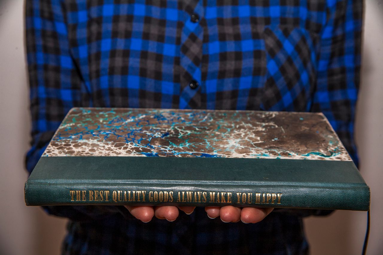 Person in blue plaid shirt holding a large hardcover book with a textured teal-and-brown cover