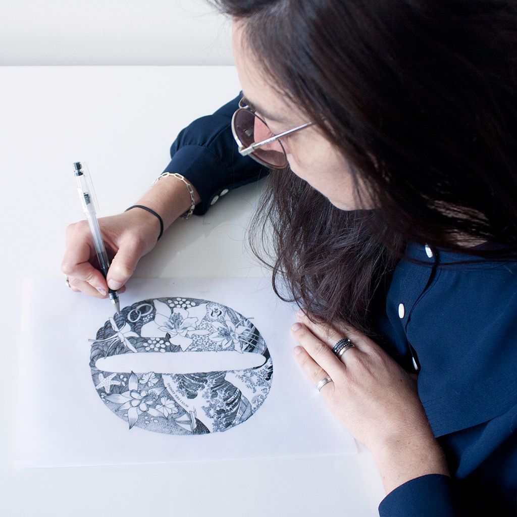 A person wearing glasses draws an intricate black-and-white circular pattern with a pen on a piece of white paper.