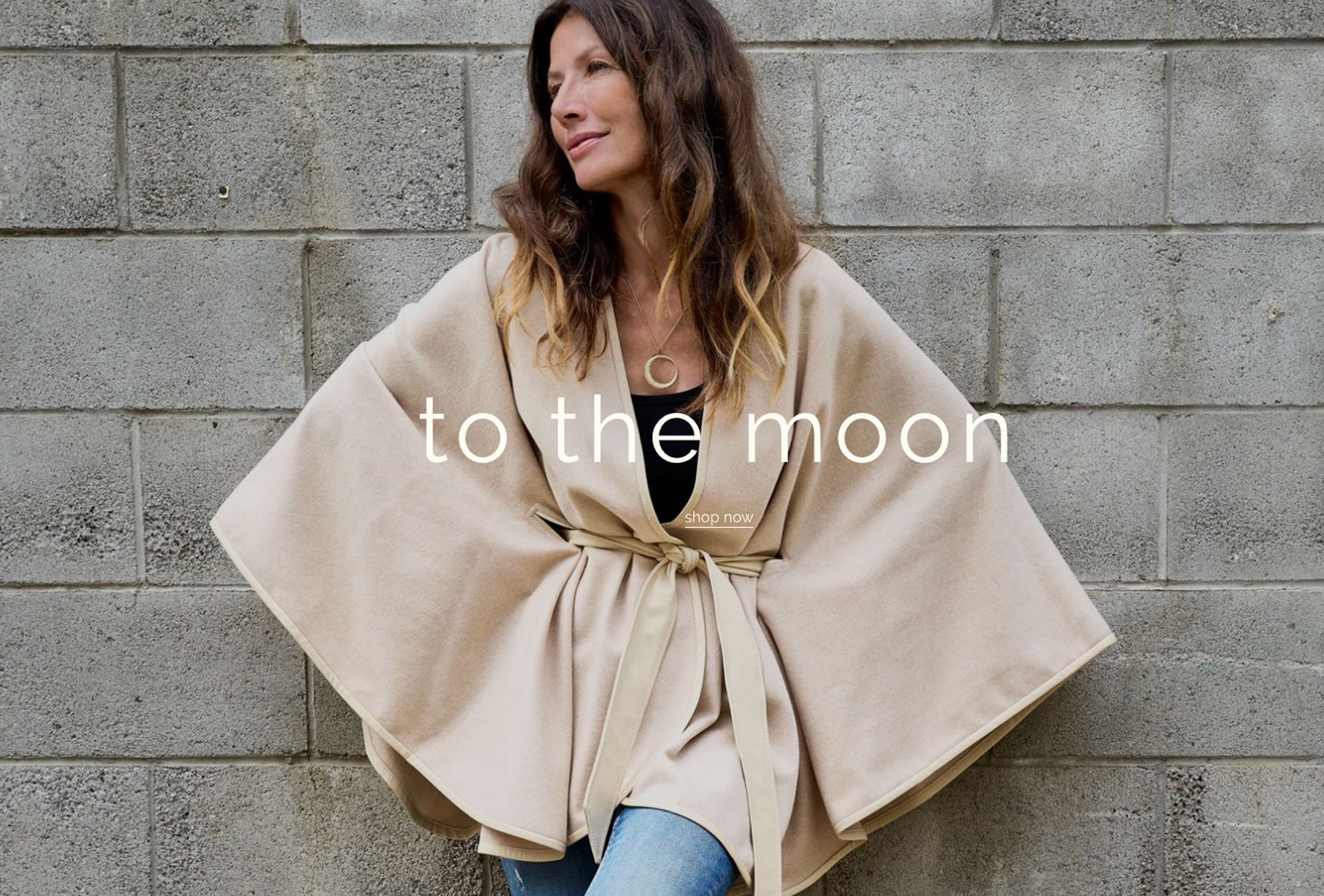 Woman in a beige wrap coat standing by a concrete wall with “to the moon” text overlaid
