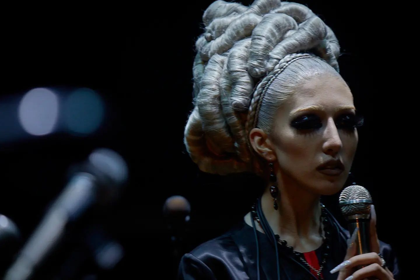 Nina Utashiro holding a microphone on a dark stage, with dramatic braided hair and blurred lights in the background