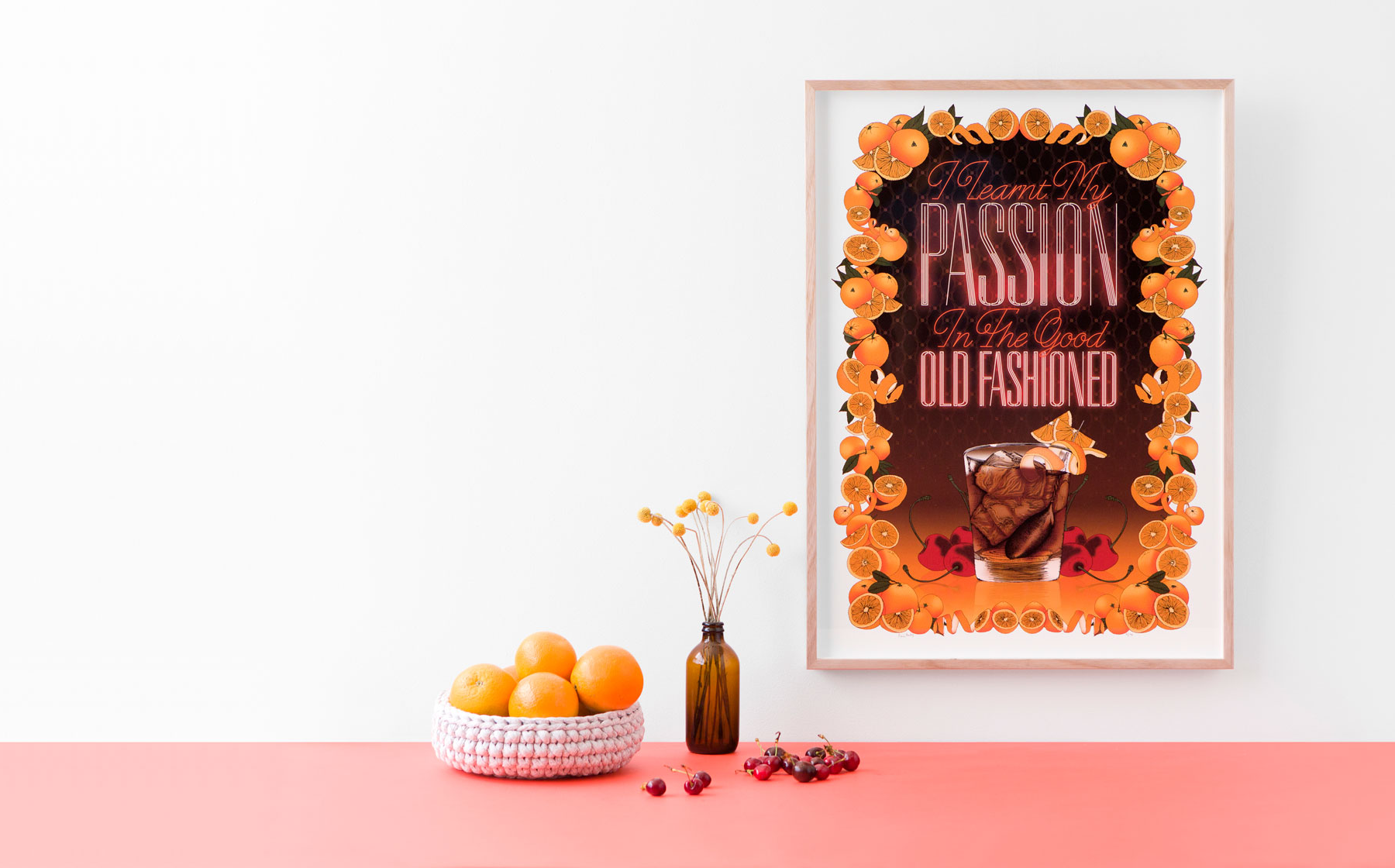 A framed art print of an Old Fashioned cocktail sits on a coral table with a bowl of oranges and a small vase of flowers.