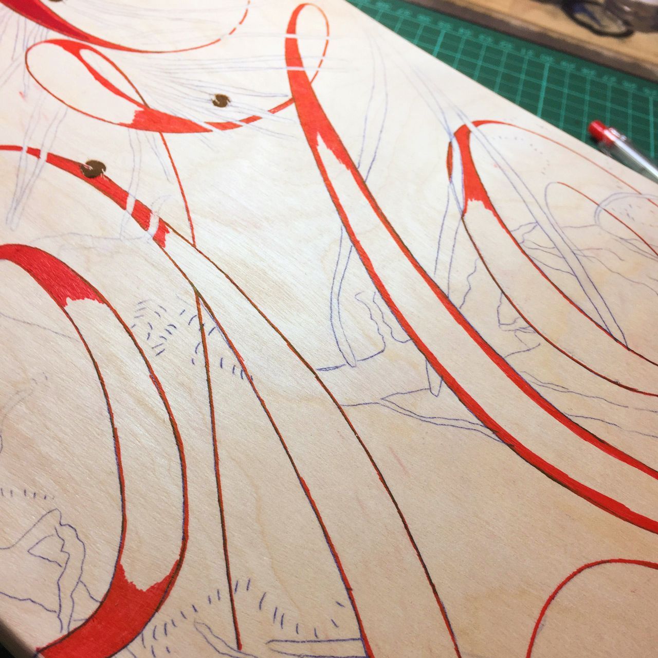A close-up of red ink swirling across a wooden surface, partially filled in with linework sketches on a craft mat.