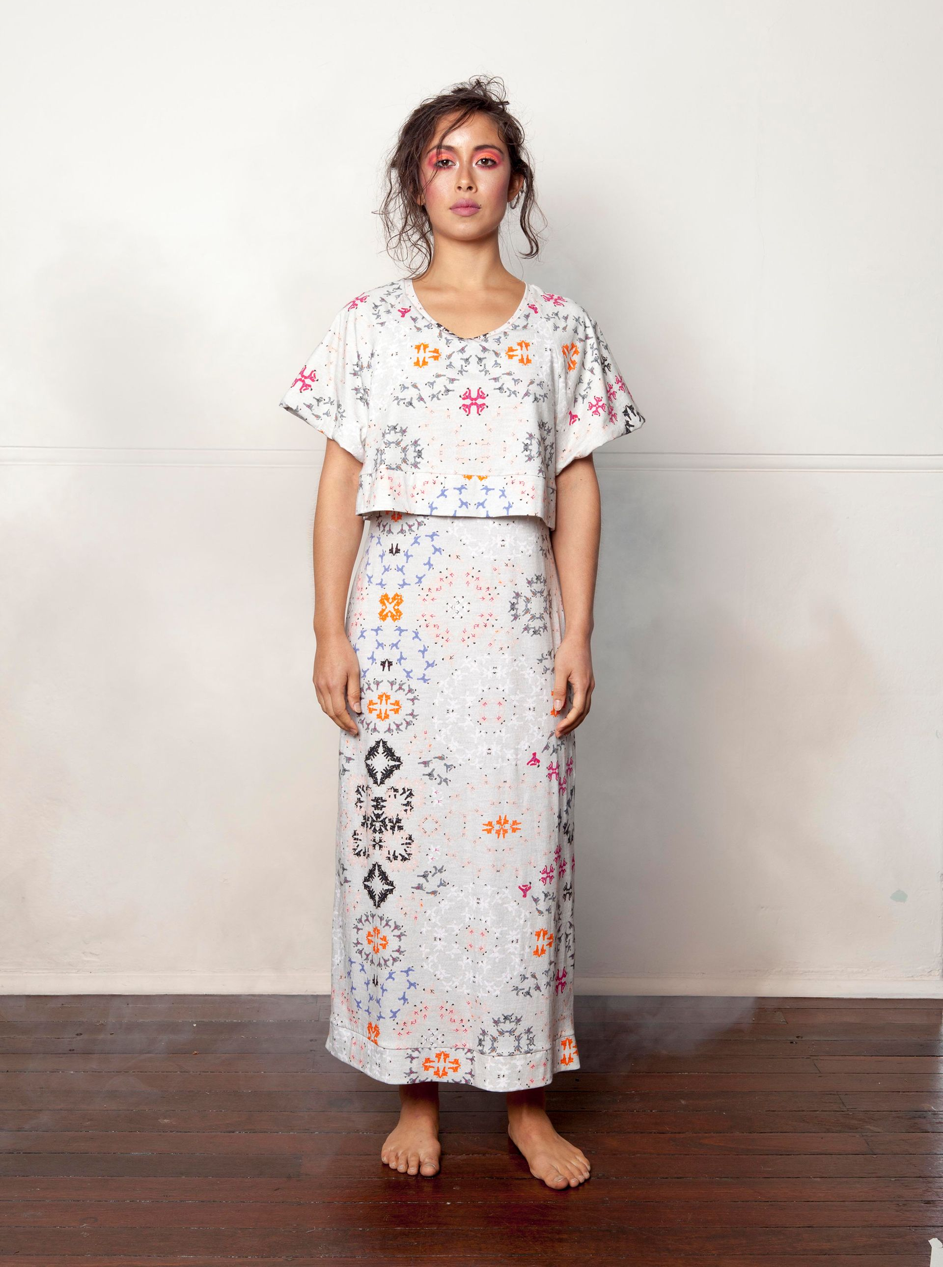 Person standing barefoot in a white patterned crop top and skirt against a plain wall