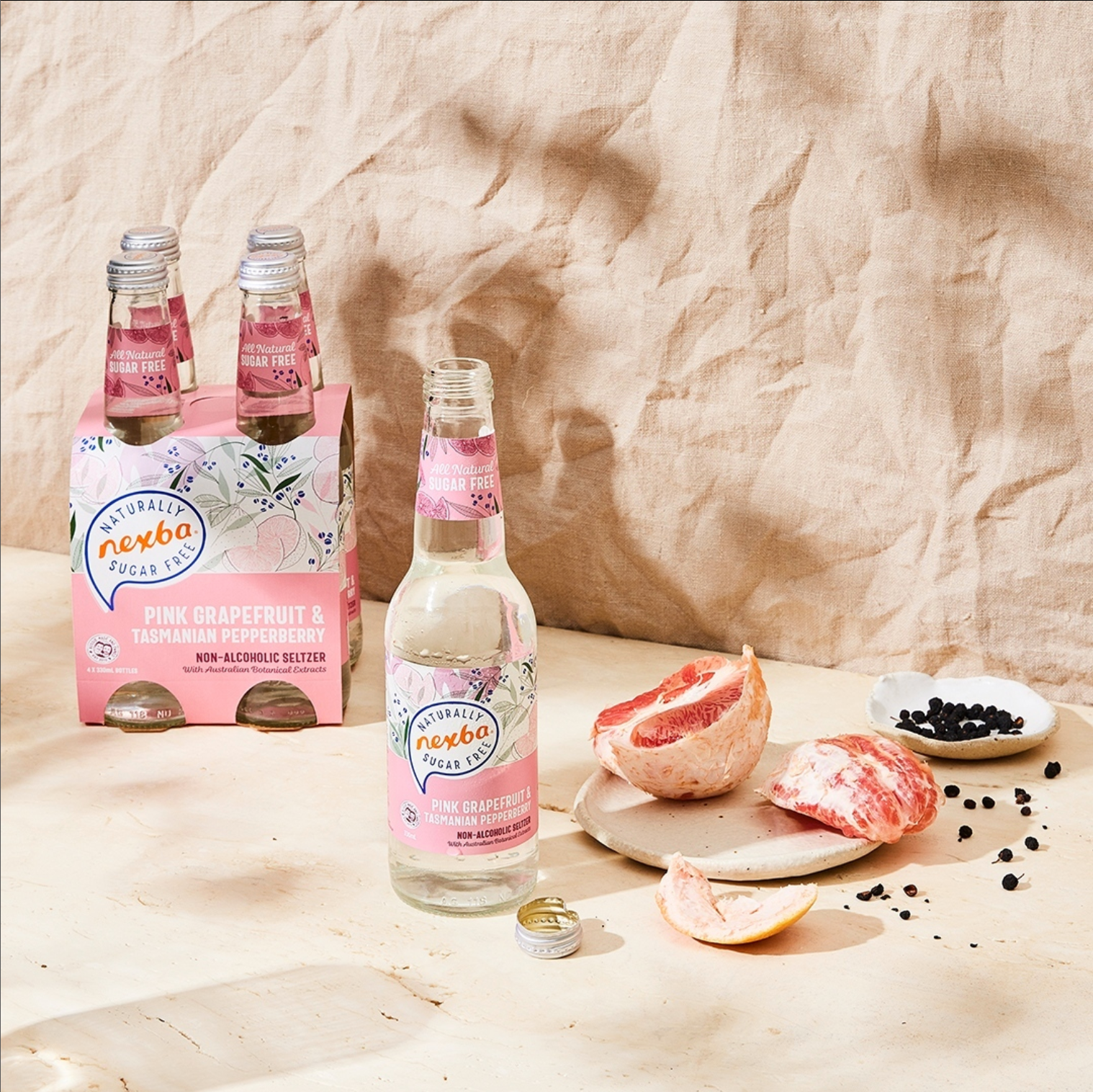 Nexba Pink Grapefruit & Juniper beverage bottles arranged with grapefruit slices and peppercorns on a textured surface.