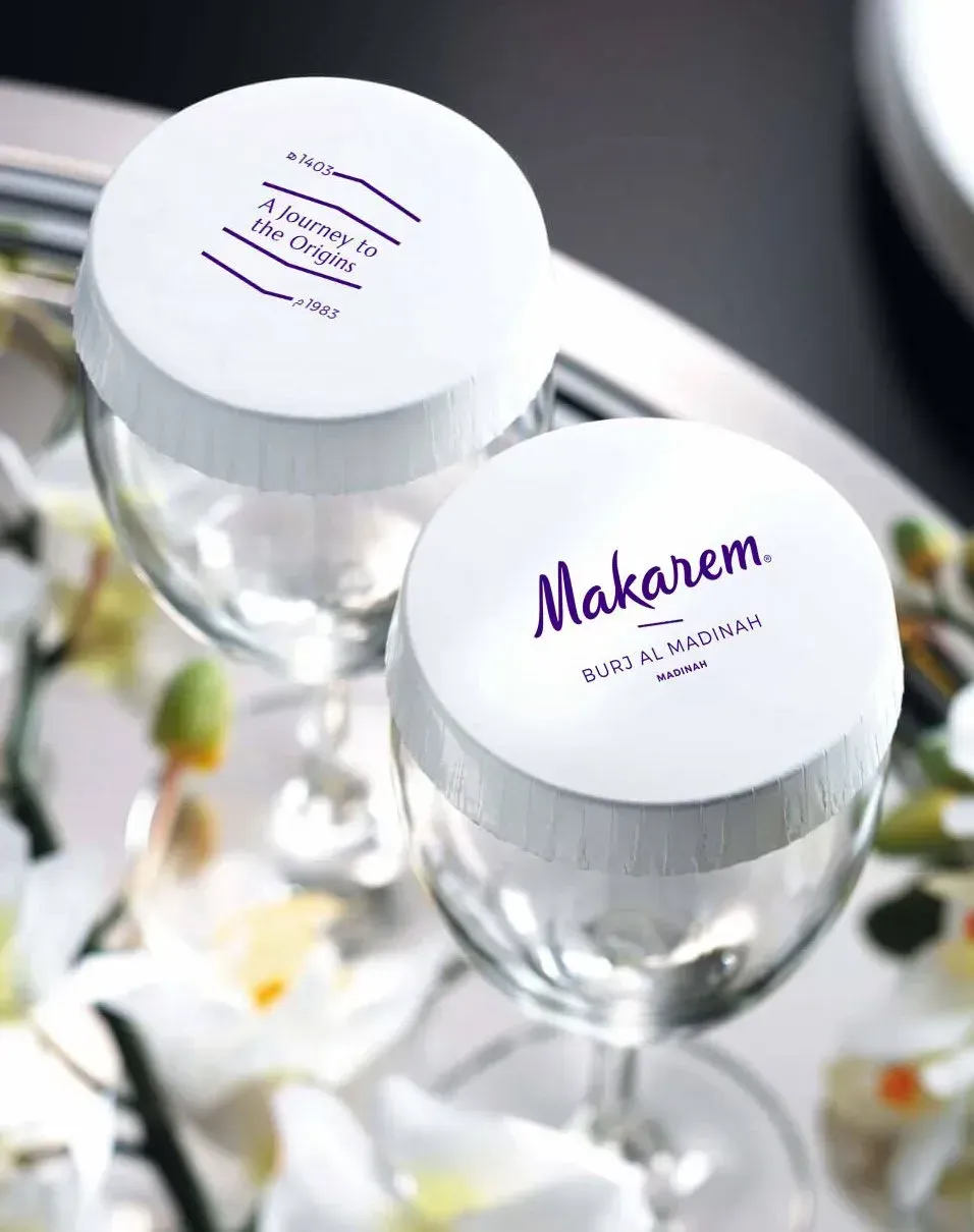 Two wine glasses with branded white lids featuring purple text, displayed among white flowers on a silver tray.
