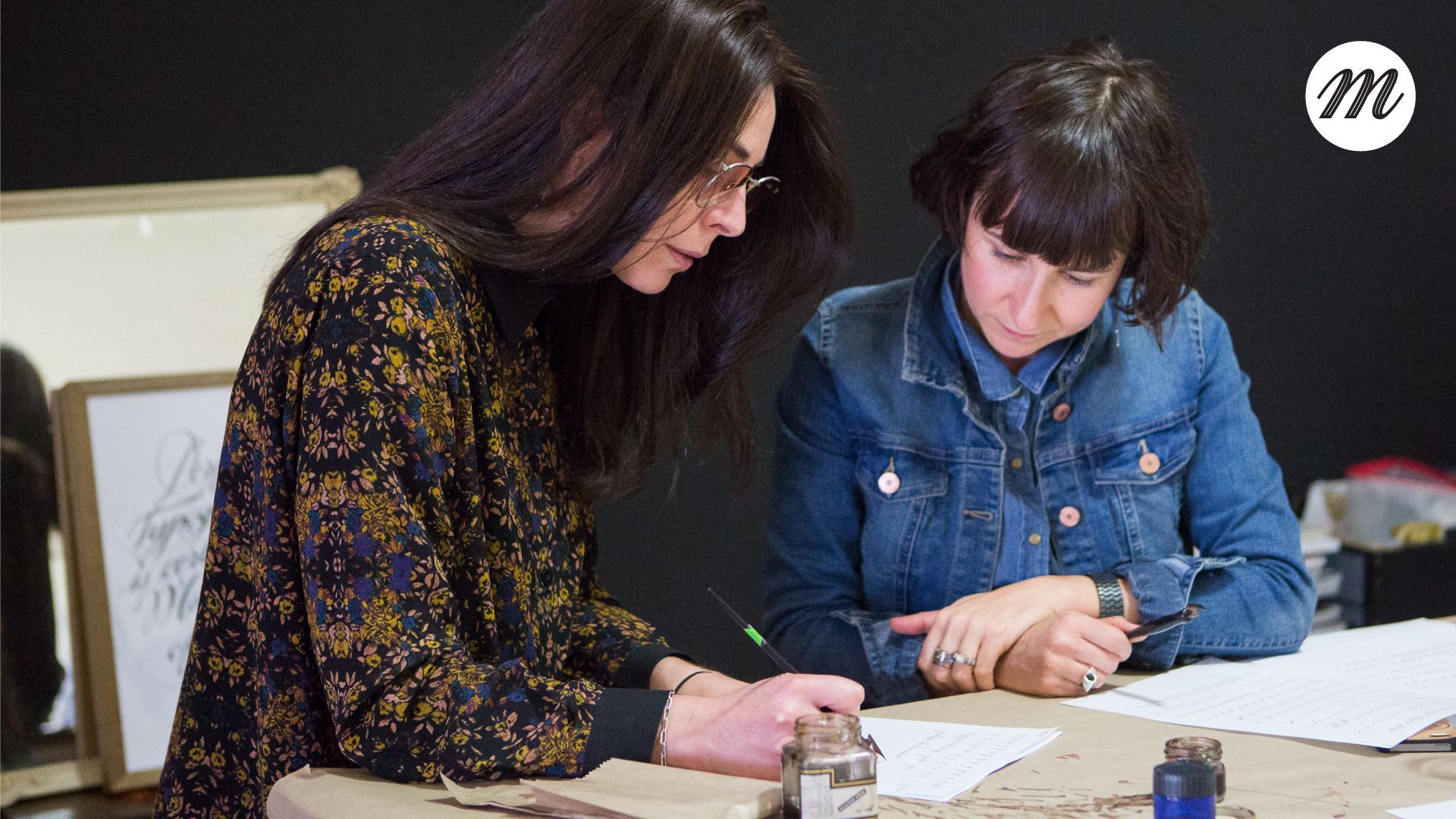 Maria Montes with a private calligraphy student leaning over a table, reviewing papers and writing with pens in a focused indoor workspace.