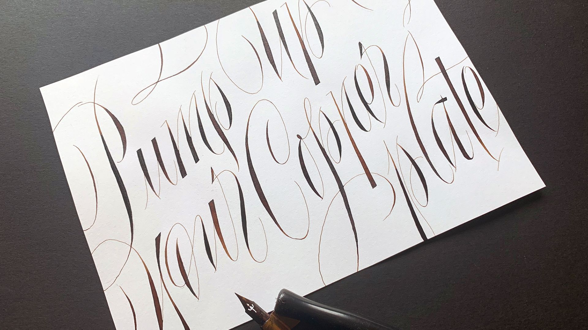 A piece of white paper with elegant, dark brown cursive calligraphy on a dark surface, accompanied by a fountain pen.