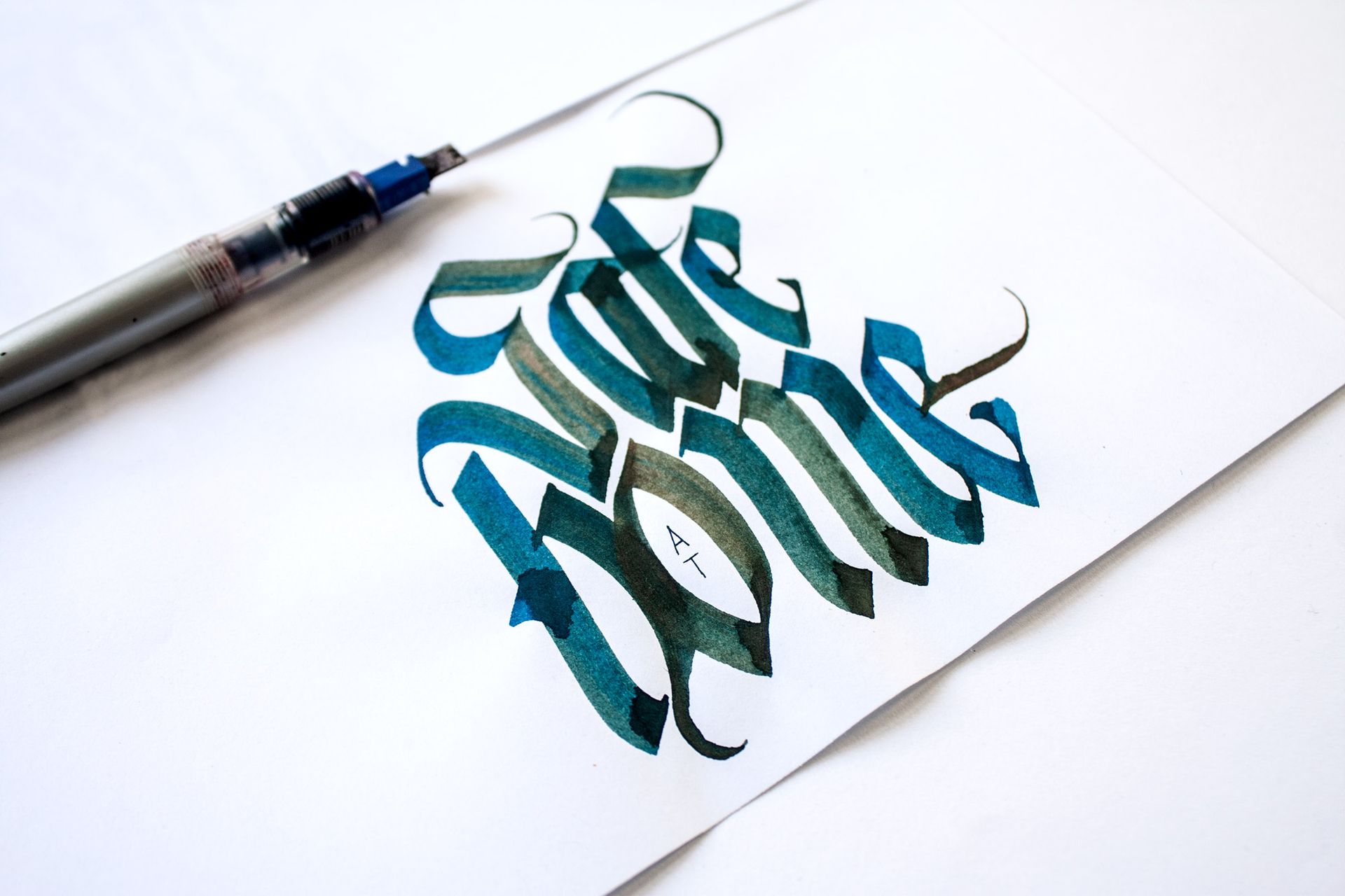 Brush lettering on white paper in teal and black ink, with a calligraphy pen nearby