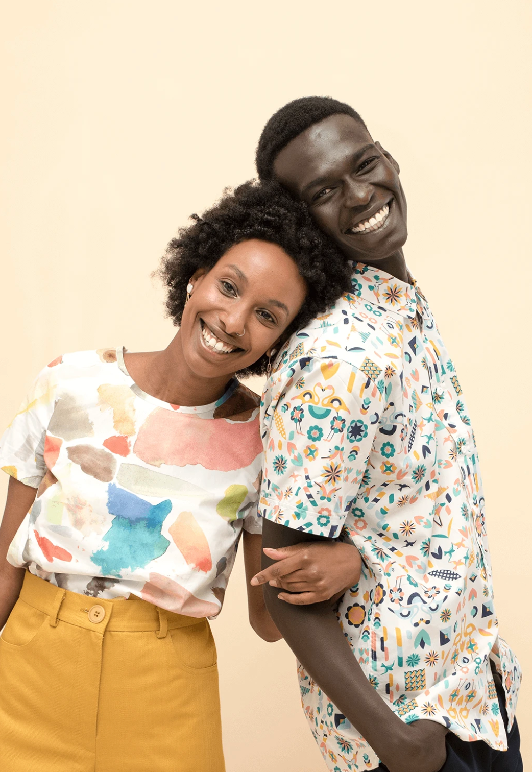 Two smiling people leaning together against a beige background, wearing colorful shirts.