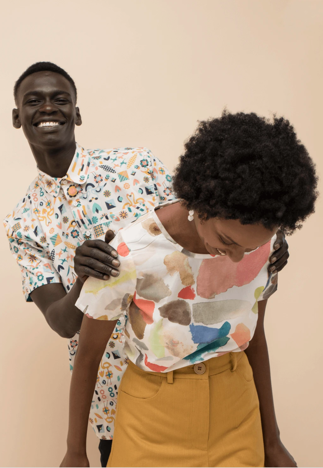 Two people smiling and posing against a plain beige wall; one leans forward in a patterned shirt and tan pants.