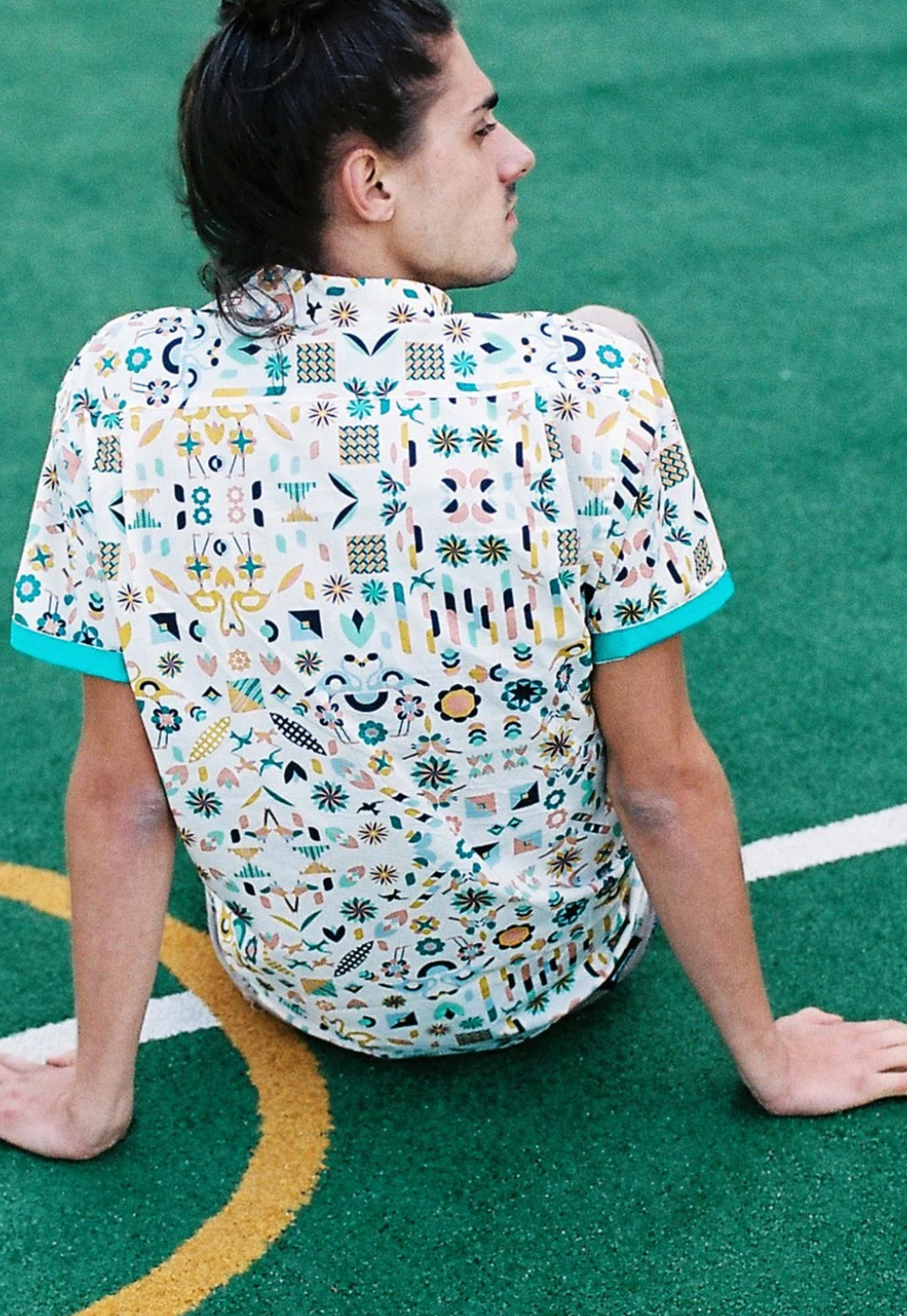Person sitting on a green sports field in a patterned shirt, leaning back and looking to the side.