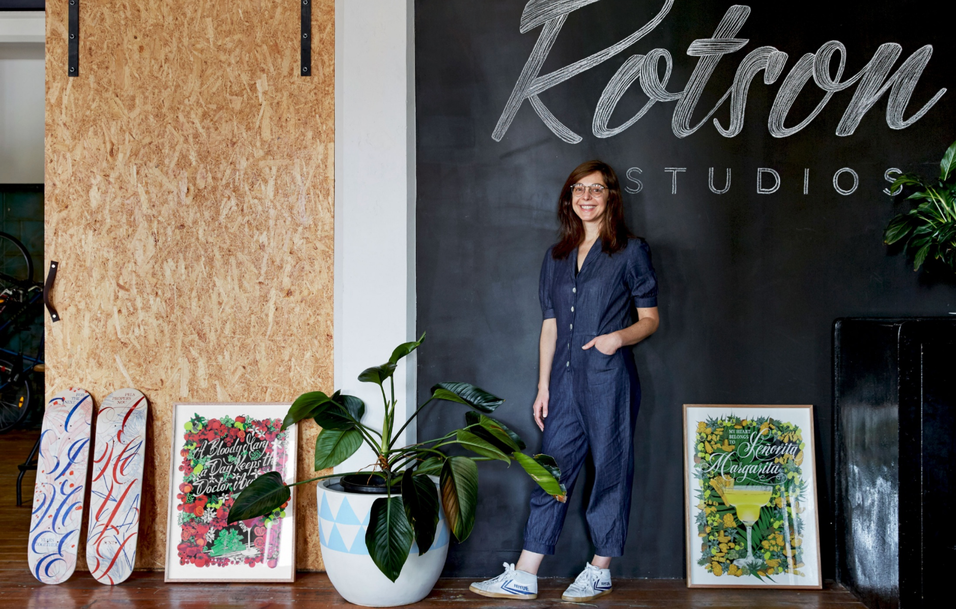 Woman standing in an art studio beside potted plants and colorful paintings on display
