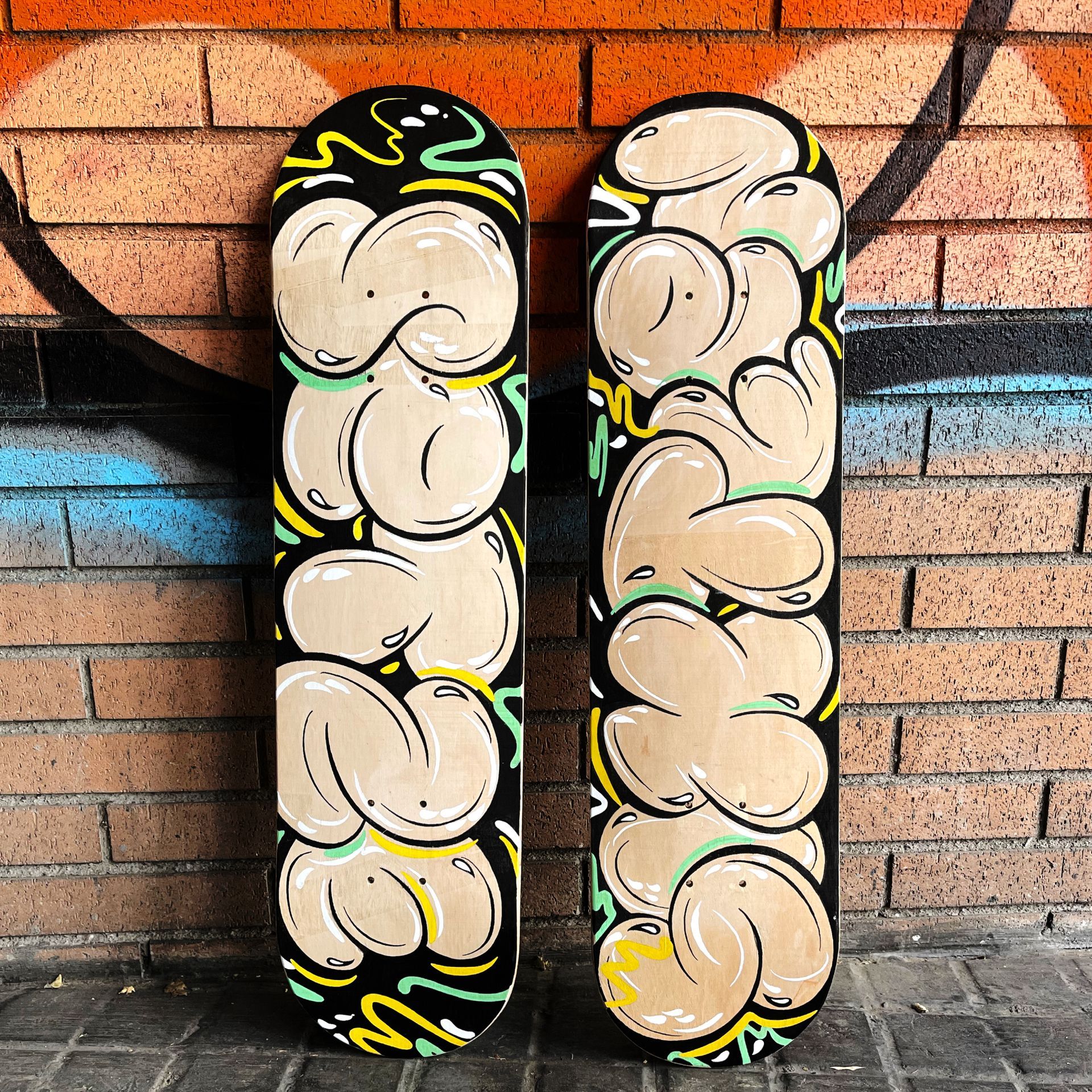 Two skateboards featuring beige, bubble-style graffiti lettering, leaning against a rustic orange brick wall.