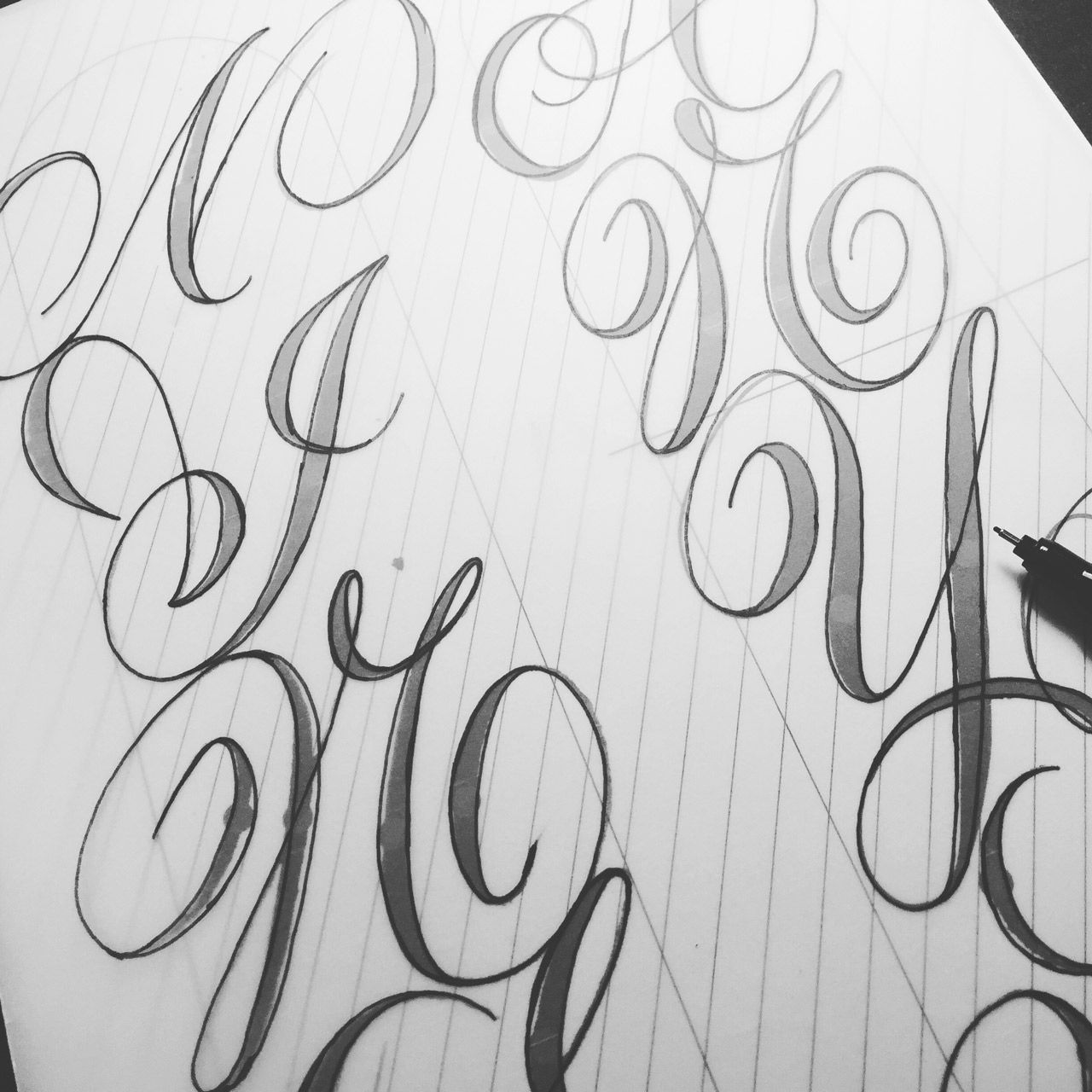 A black and white image shows a hand-drawn calligraphy sketch on grid paper with a pen resting on the side.