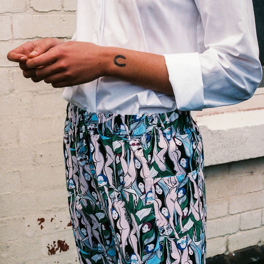 Person in a white shirt and patterned pants, arms crossed, standing against a white brick wall.