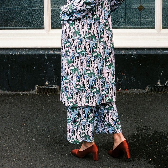 Person in a floral patterned outfit and red heels standing on a wet sidewalk by a window.
