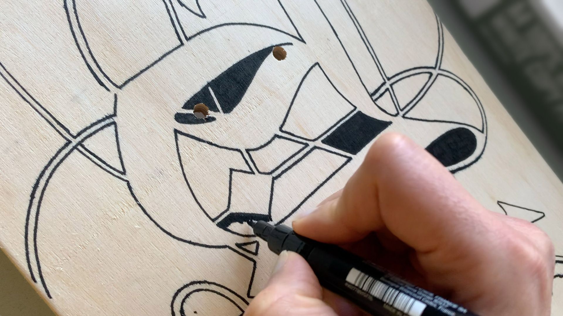 A hand uses a black marker to color in the geometric lines of a stencil-like illustration on a light-colored surface.