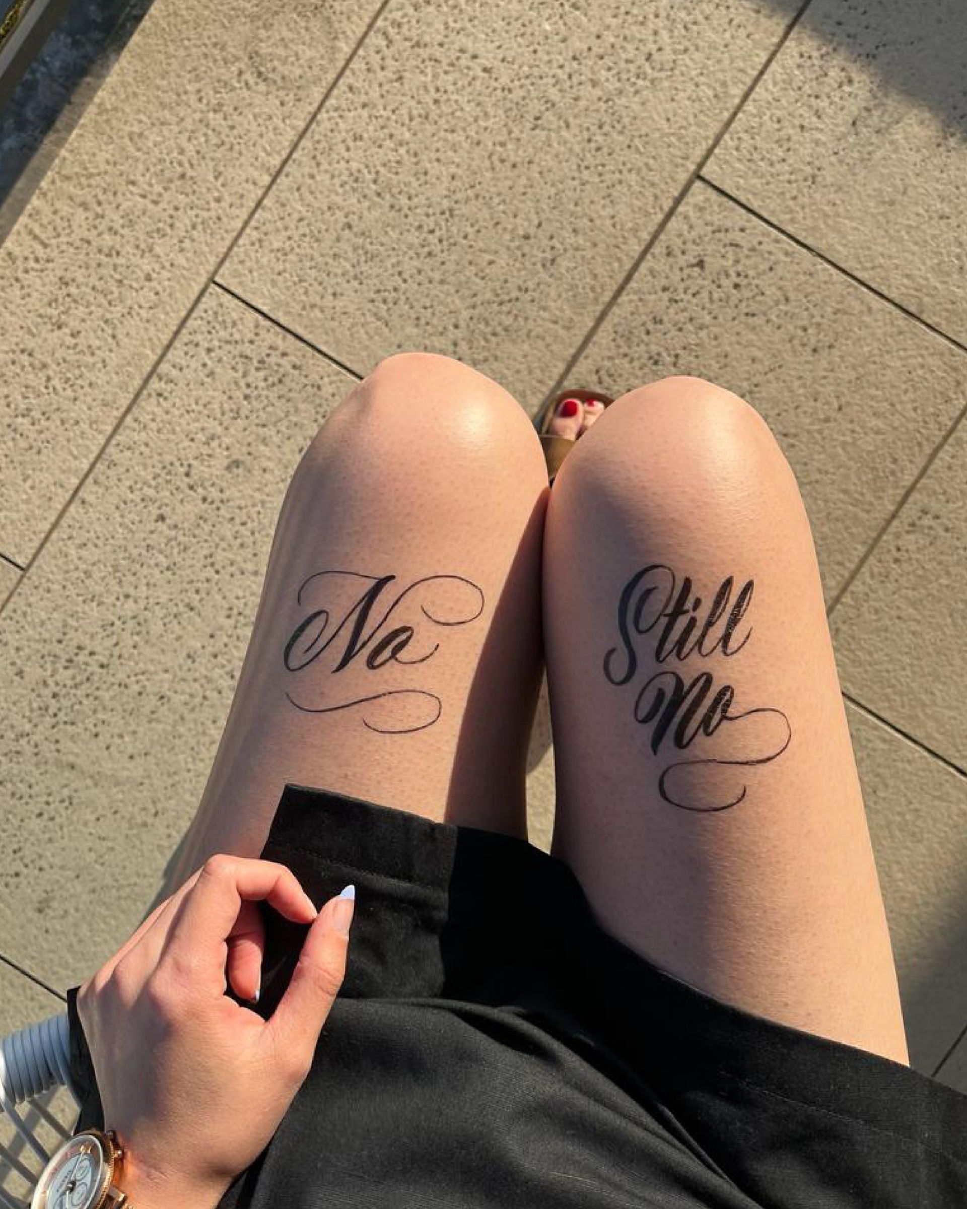Legs in black shorts showing matching thigh tattoos reading “No” and “Still No” on a tiled floor.