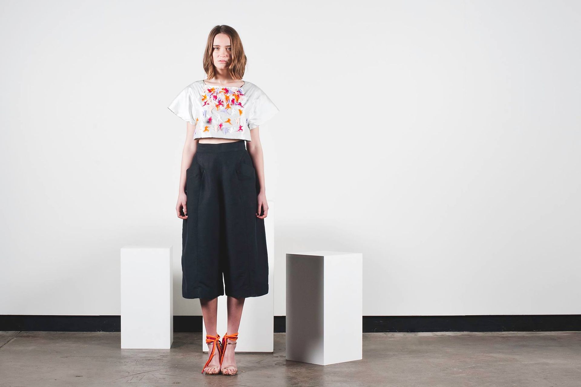 Person standing in a white room, wearing a white graphic top, black wide-leg pants, and red sandals.
