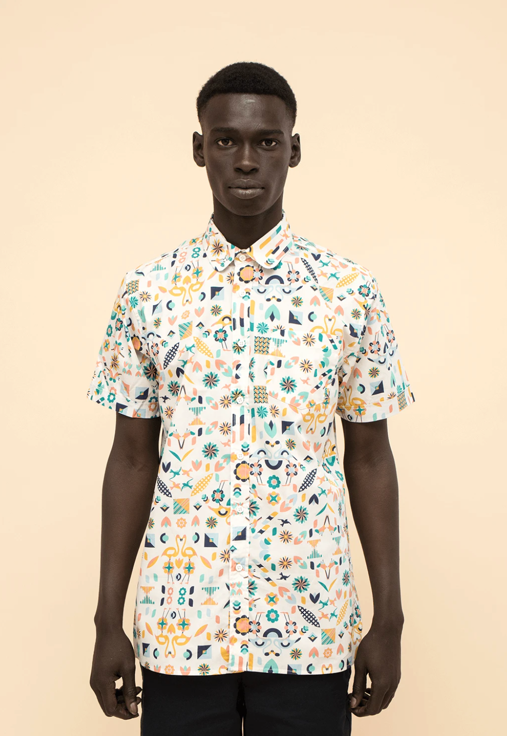 Person standing against a beige background wearing a white patterned short-sleeve shirt.