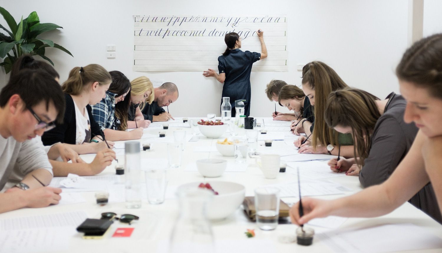 People seated at a long table in a workshop, writing while a presenter writes on a whiteboard.