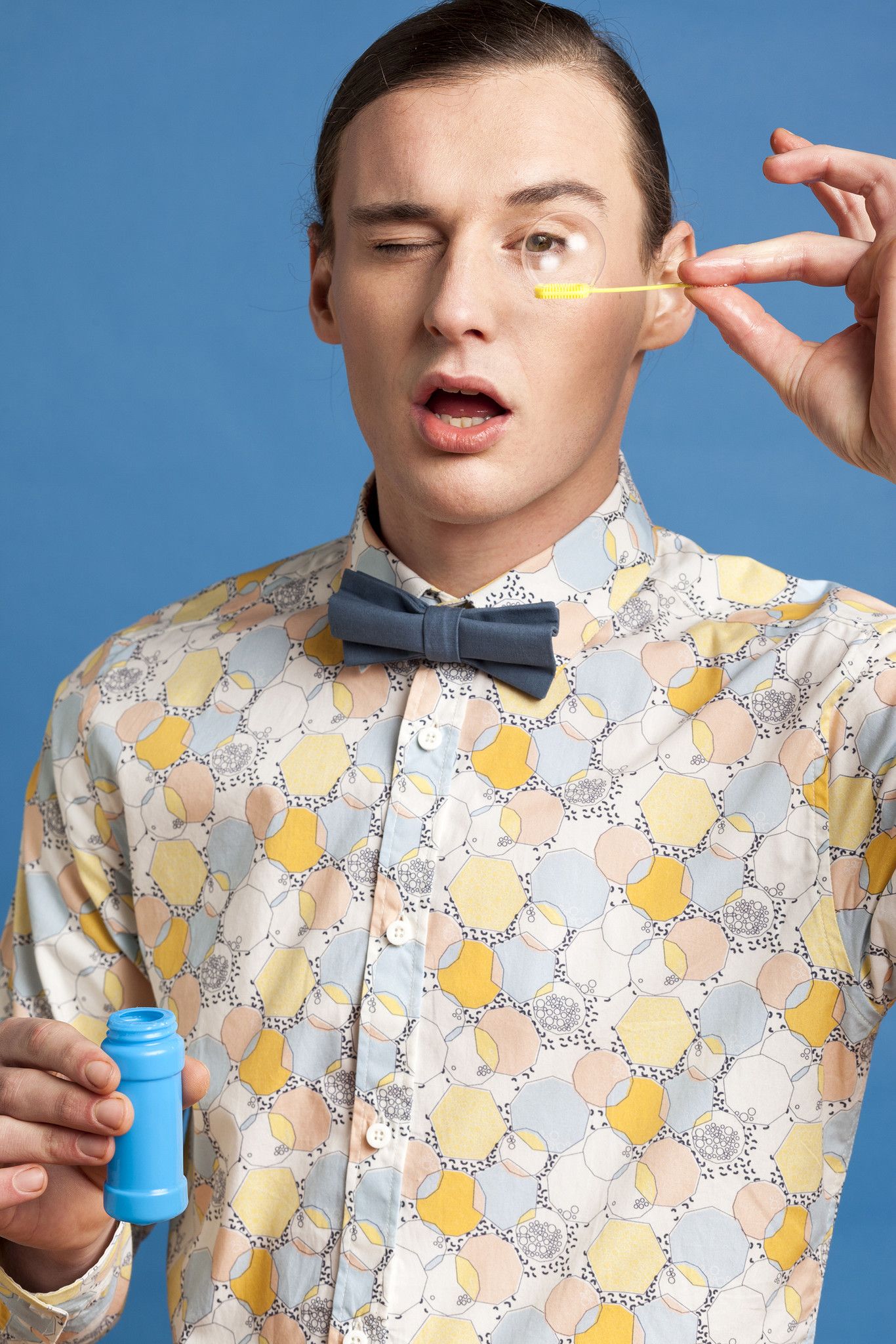 Man closing one eye while applying eye makeup with a brush, holding a blue container against a blue background