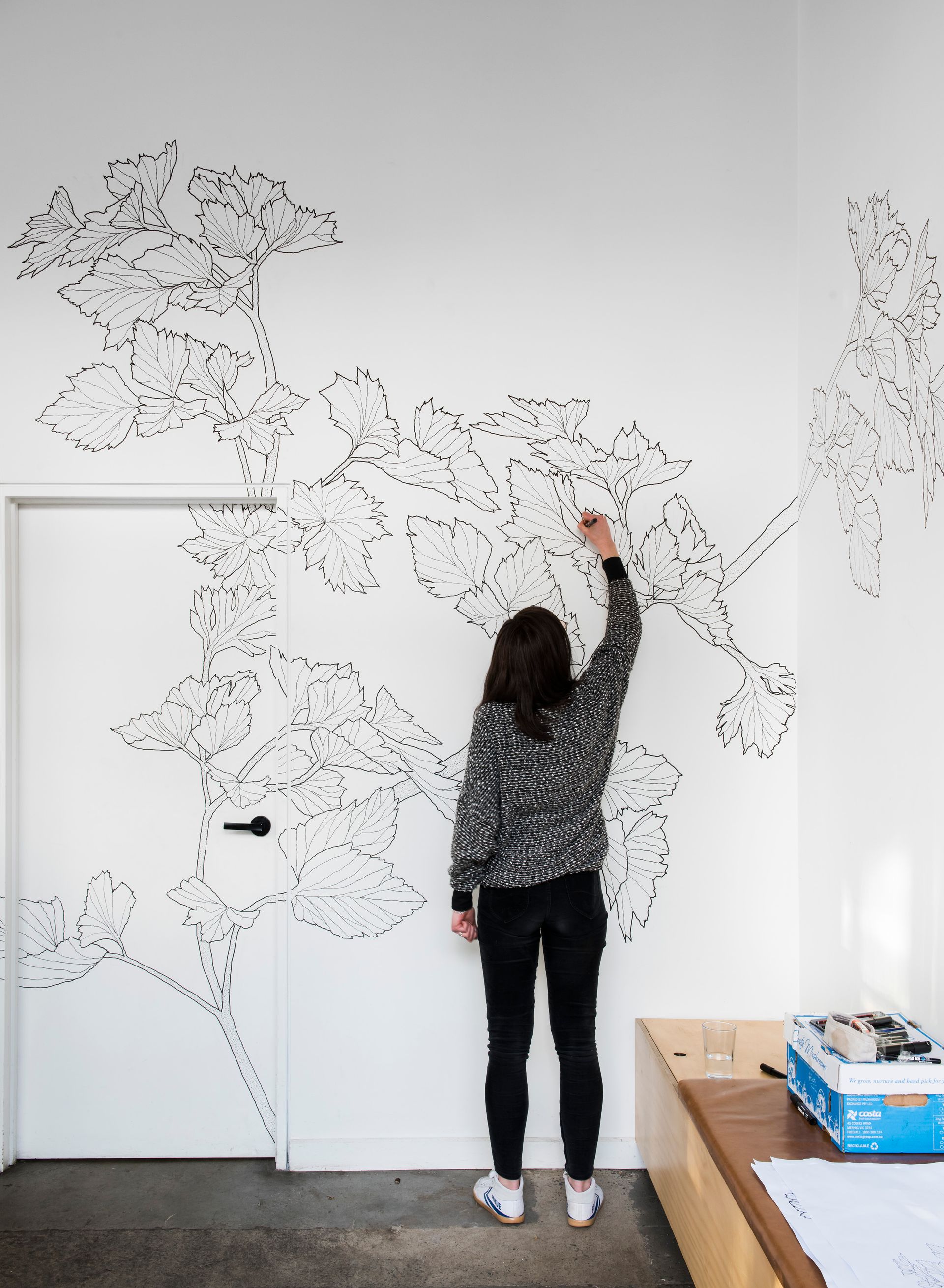 Person drawing on a white wall covered with a black line-art mural in a bright room