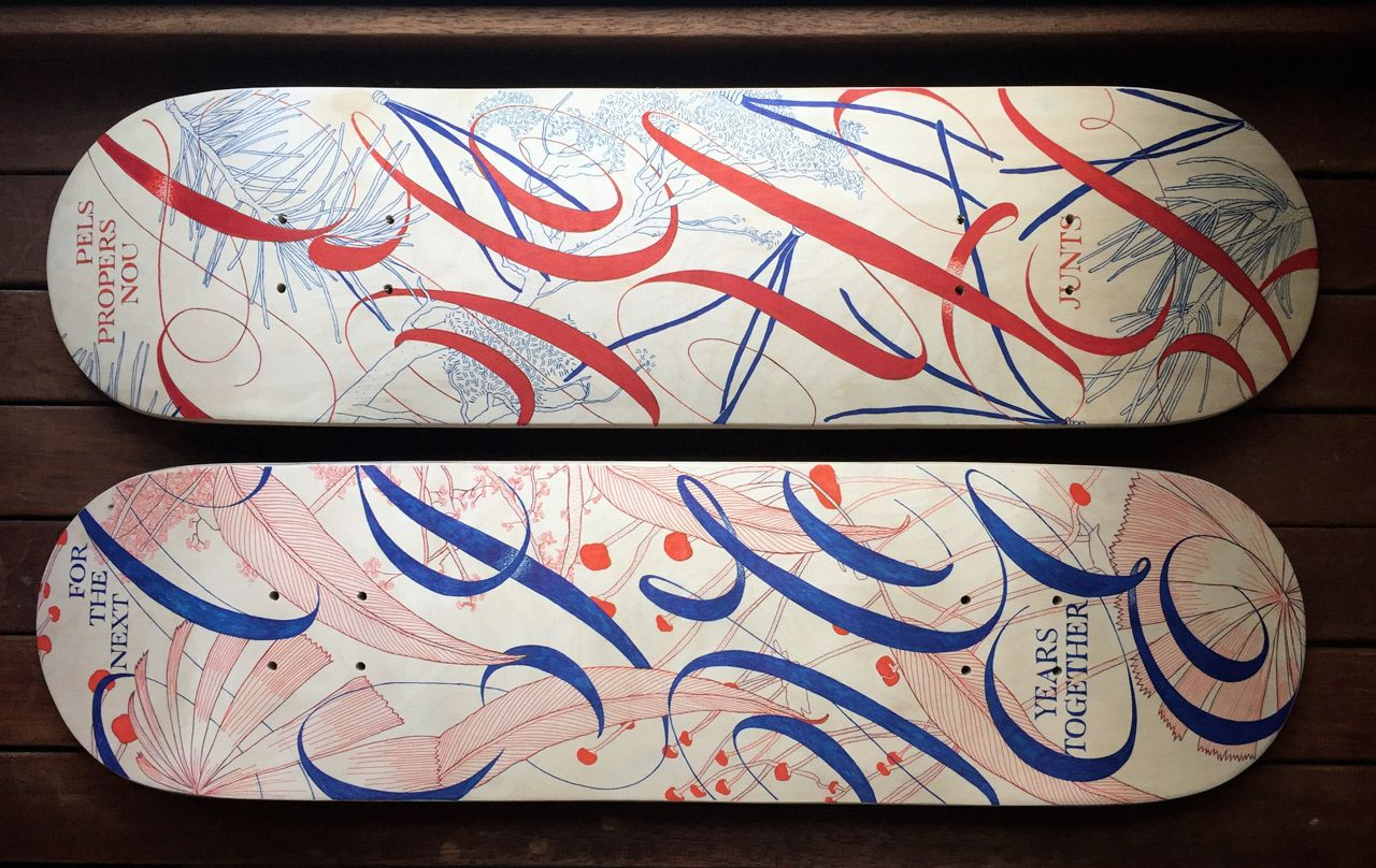Two skateboard decks with white backgrounds and intricate red and blue abstract calligraphy designs, resting on wood.