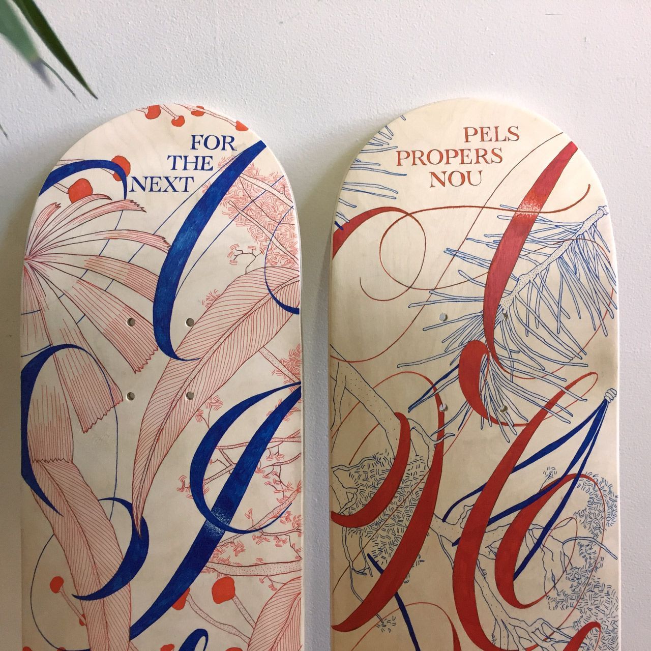 Two skateboard decks featuring ornate, stylized calligraphy in red and blue against a cream background.