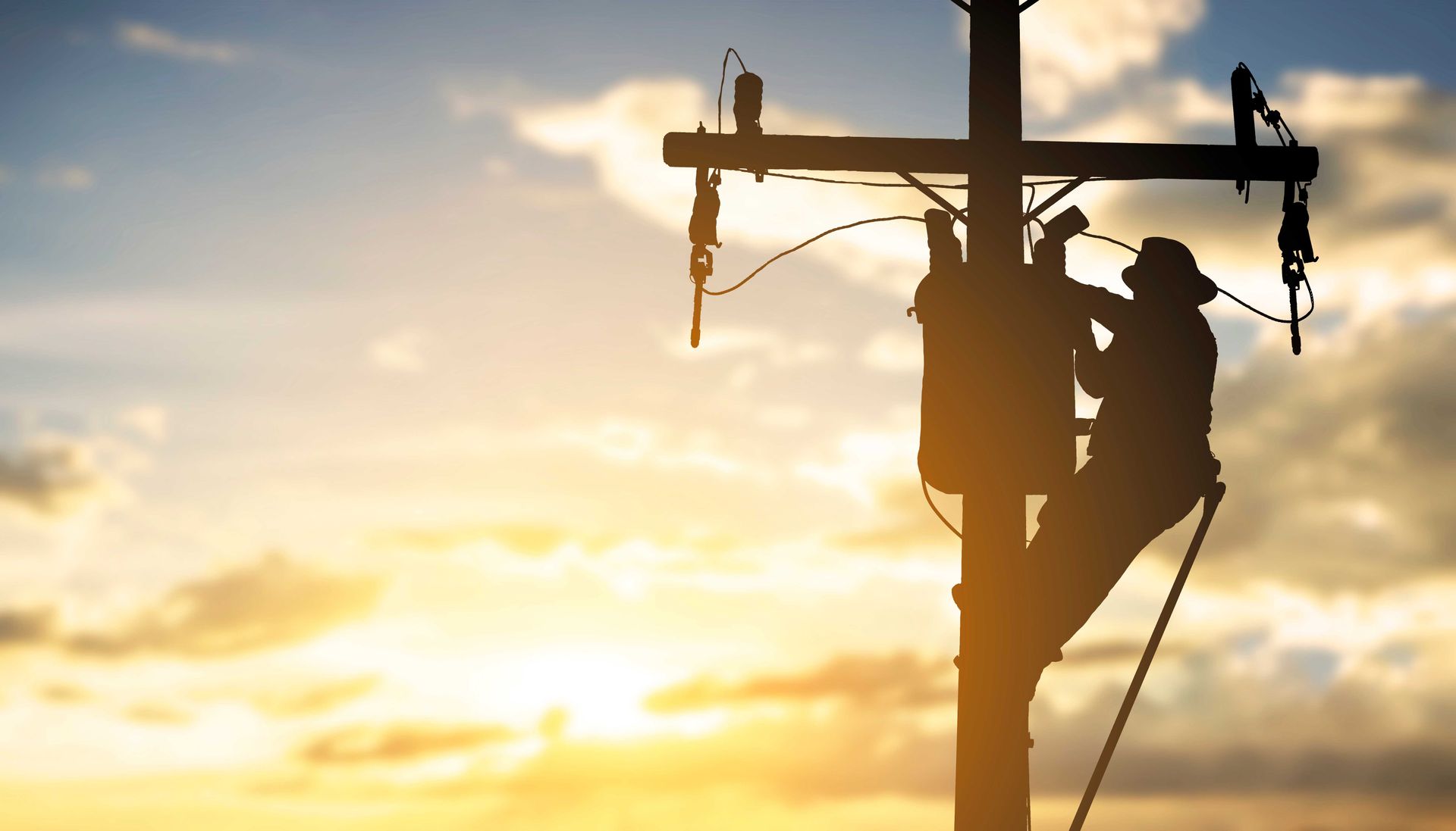 Silhouette d'une personne travaillant sur un poteau électrique, sur fond de coucher de soleil.