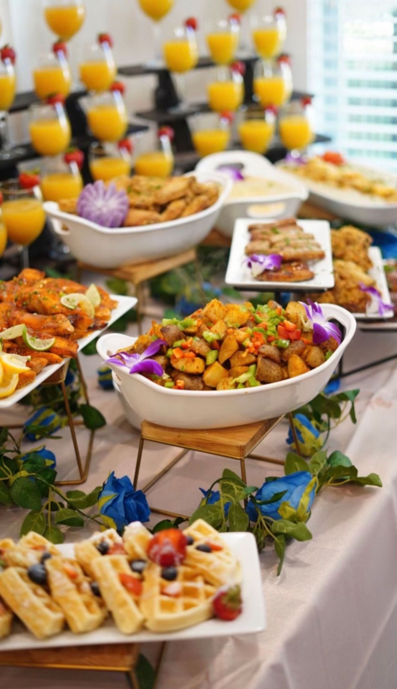 Brunch buffet with waffles, fried food, and stacked orange juice glasses, on a table with floral decorations.