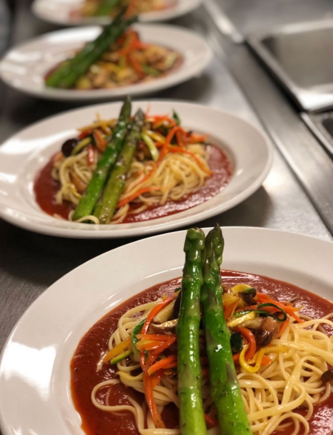 Plates of pasta with tomato sauce, vegetables, and asparagus, lined up on a stainless steel surface.
