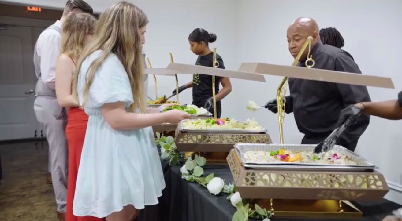People at a buffet, served by staff. Food trays on a decorated table.