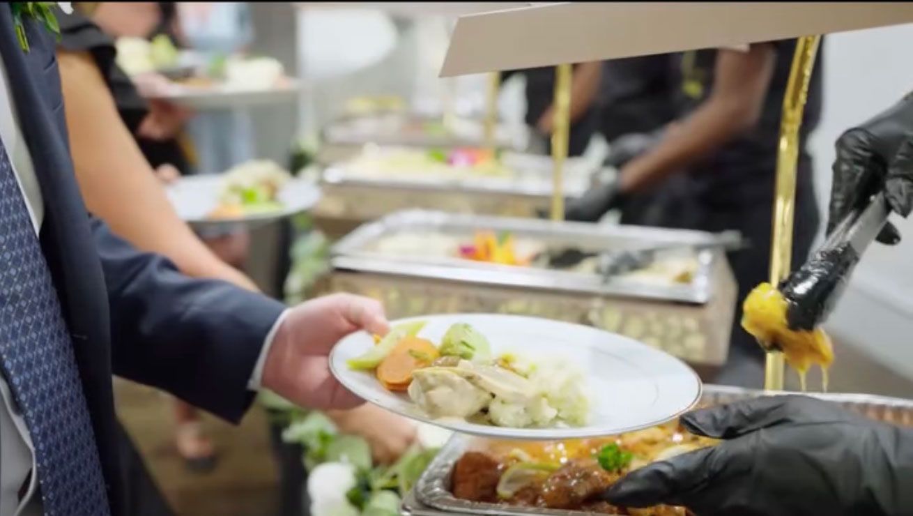 Guests at a catered event, served food from a buffet. Person in suit receives a plate. Staff wearing black gloves.