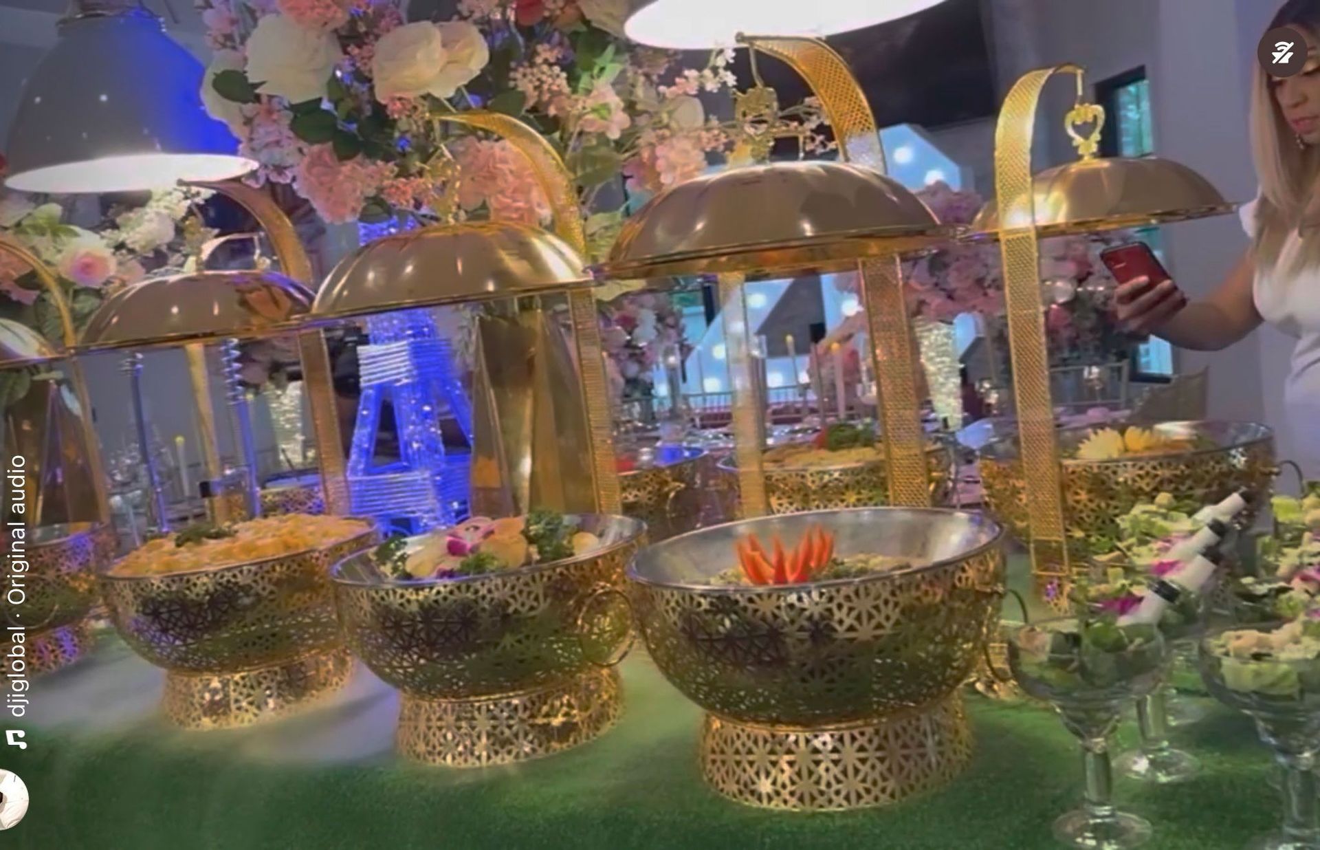 Golden food displays with food and flowers at a party. A woman in a white dress is nearby.