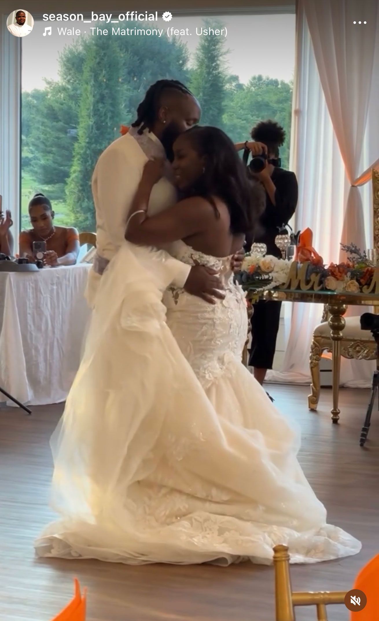 Bride and groom dance at reception; she wears white gown, he a white suit. Green trees visible in background.