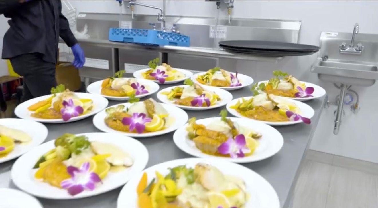Plated meals on a stainless steel counter, prepared in a commercial kitchen setting. A person in black uniform is working.