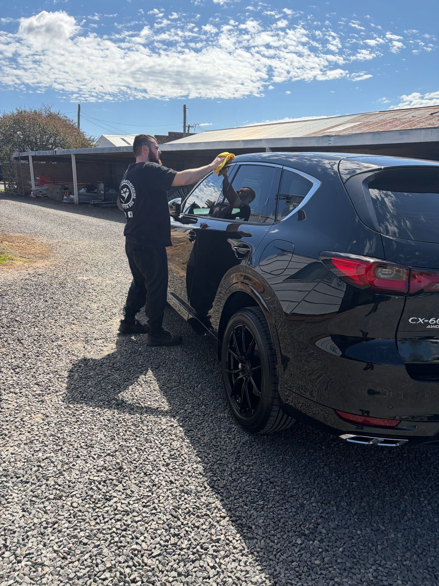 Person washing a black SUV outdoors on a gravel driveway under a bright blue sky