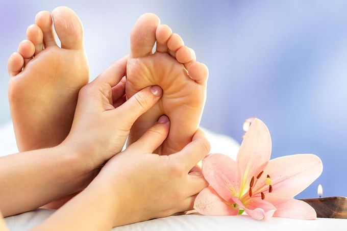 Primo piano di una persona che riceve un massaggio ai piedi, con un fiore rosa appoggiato nelle vicinanze su una superficie morbida e chiara.