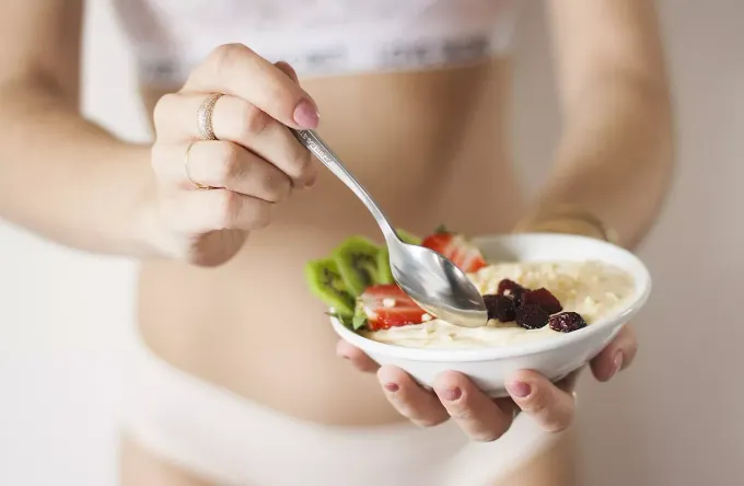 Una persona tiene in mano una ciotola bianca piena di farina d'avena, fragole a fette, kiwi e frutta secca, pronta per essere mangiata con un cucchiaio.