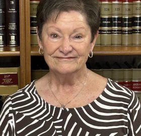 Woman with short brown hair in front of a bookshelf wearing a patterned shirt and a necklace, smiling.