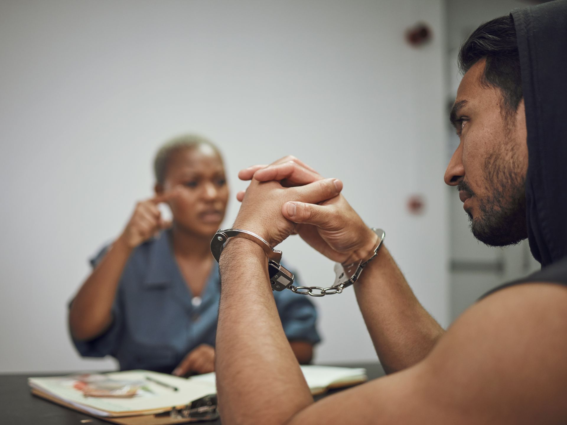 Man in handcuffs at table in interrogation room; investigator gestures.