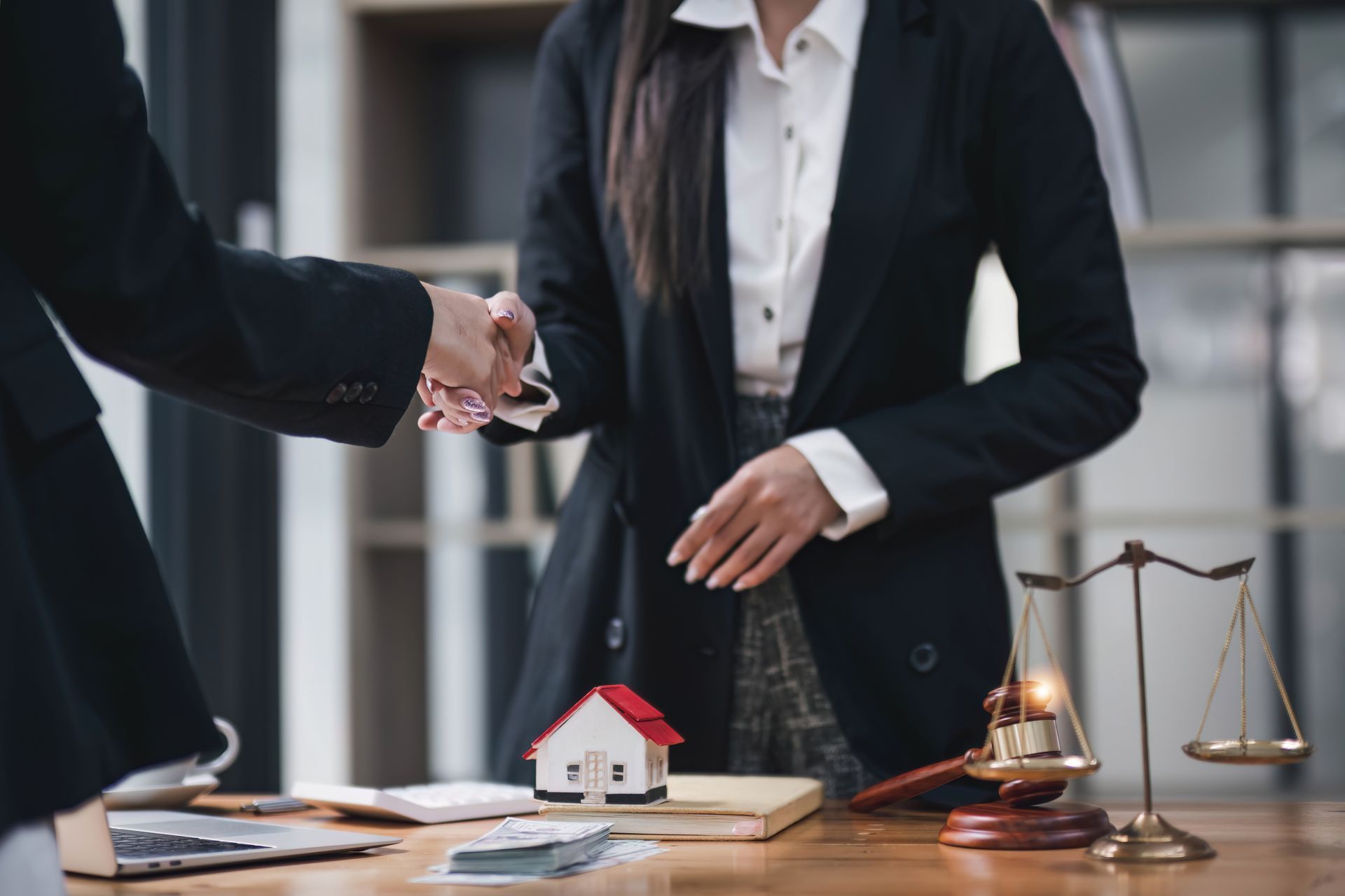 Two people shaking hands near a gavel, scales, and a house model on a desk.