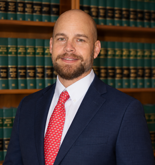 Man in suit, red tie, beard; standing in a library with bookshelves of books in the background.